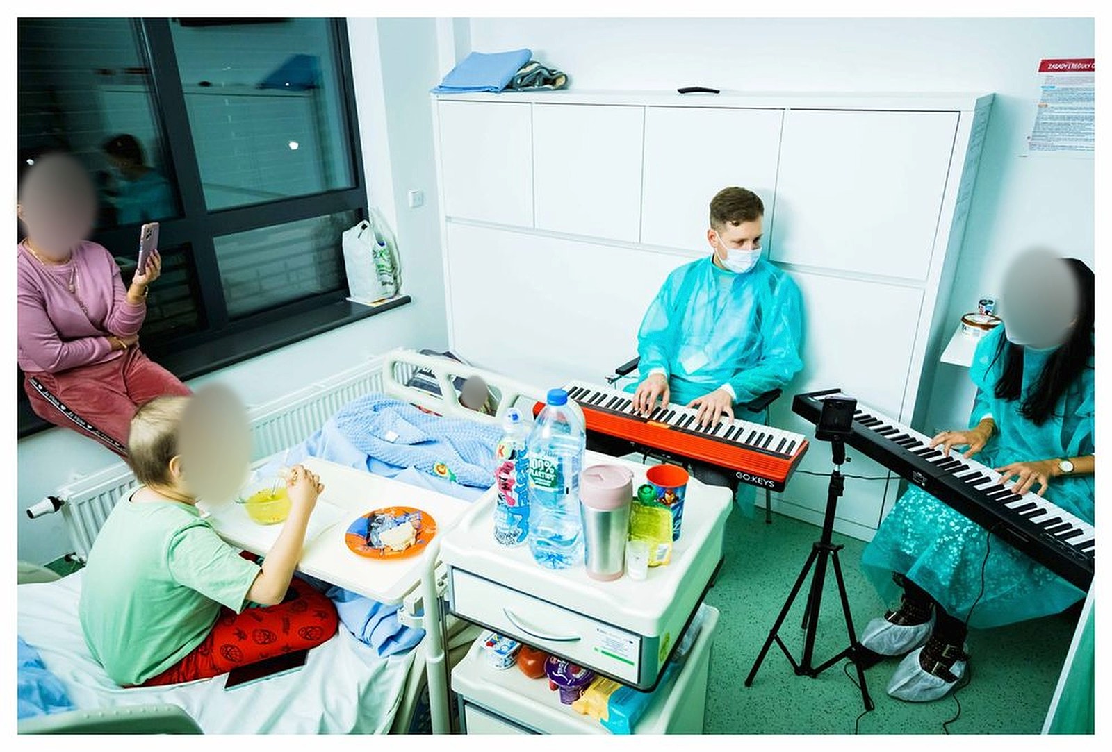 Sala szpitalna: dwoje muzyków w fartuchach gra na keyboardach przy łóżku dziecka, obok dzieci i rodzic; twarze osób trzecich zasłonięte
