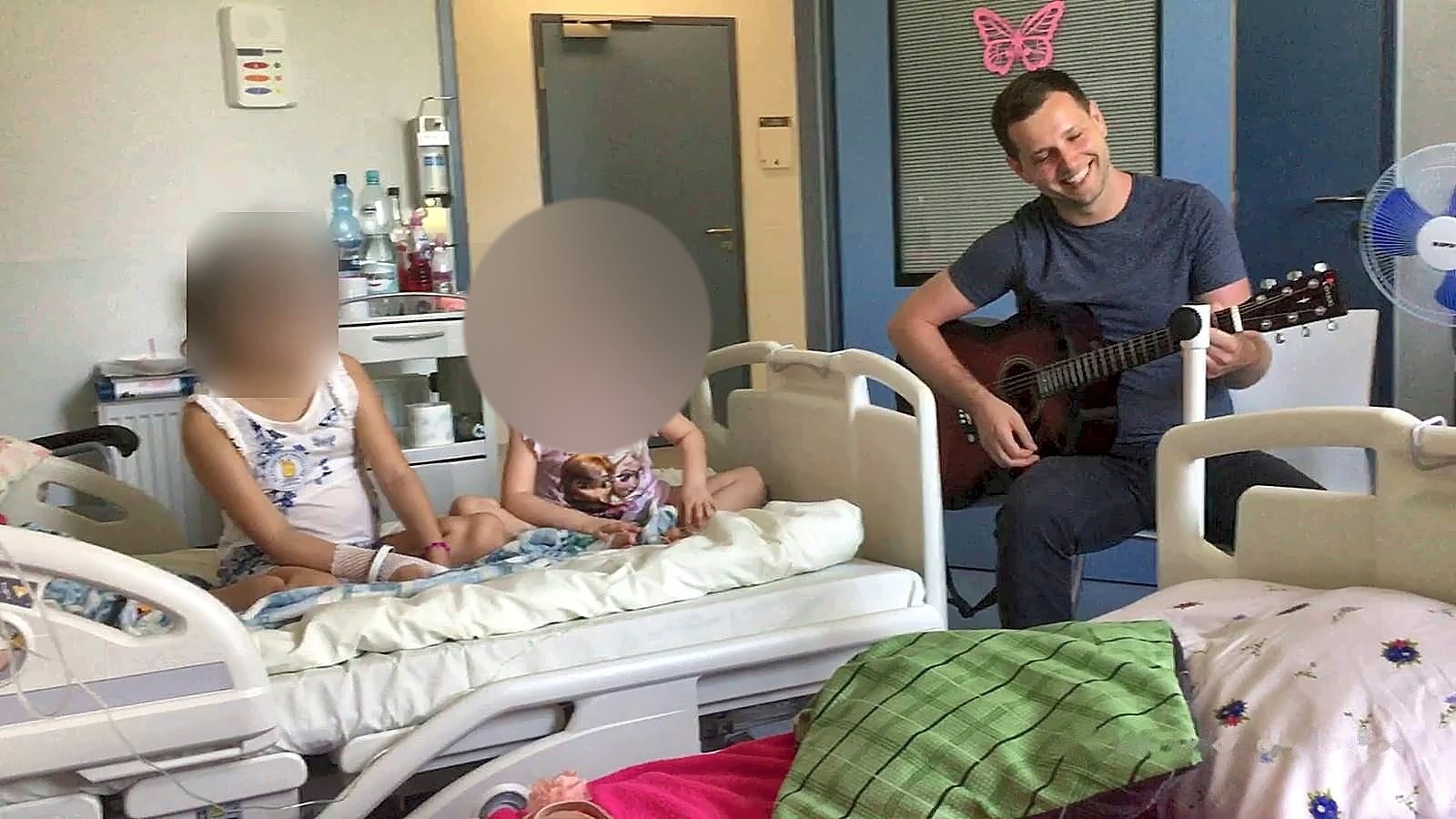 Sala szpitalna: Michał Drezner gra na gitarze, dwie dziewczynki słuchają z łóżek; twarze dziewczynek zasłonięte