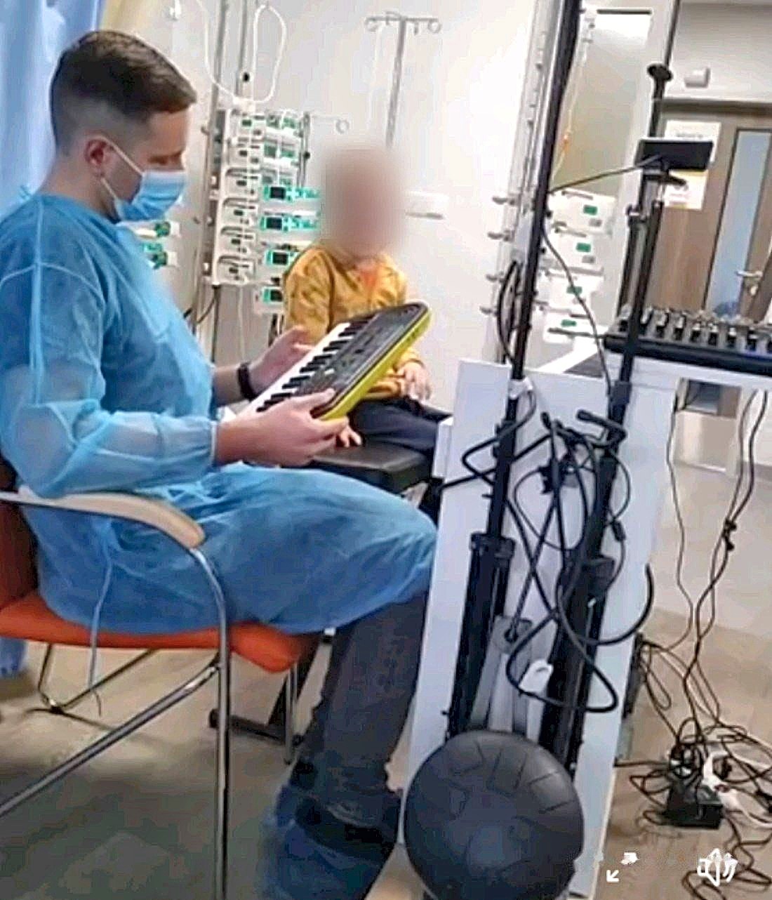 Sala szpitalna: Michał Drezner w niebieskim fartuchu i maseczce trzyma na kolanach żółty keyboard, obok siedzi dziecko (twarz zasłonięta)