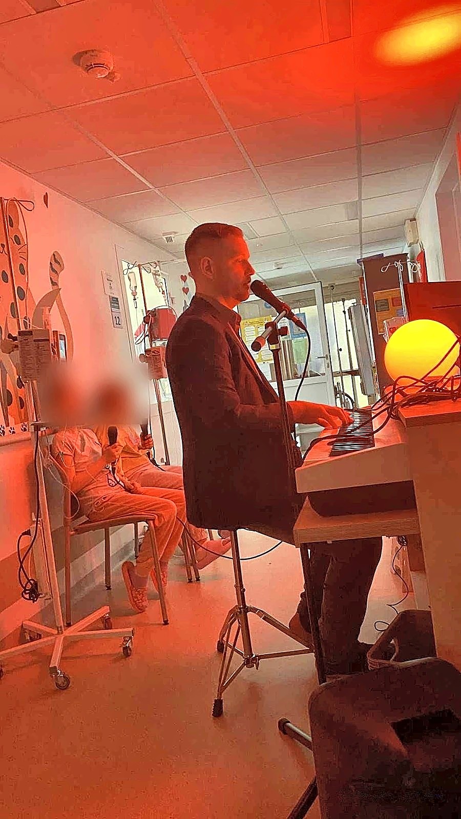 Michał Drezner śpiewa przy keyboardzie w sali szpitalnej w ciepłym świetle, obok dzieci z mikrofonami (twarze zasłonięte)