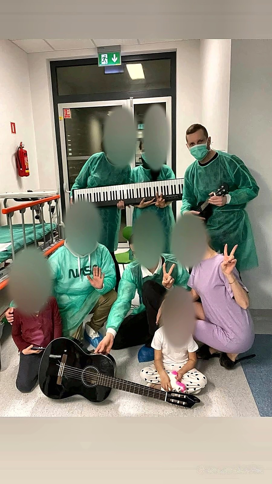 Korytarz szpitalny: kilkoro osób w fartuchach ochronnych i maseczkach z keyboardem i gitarą, Michał Drezner z ukulele po prawej, pozostałe twarze zasłonięte