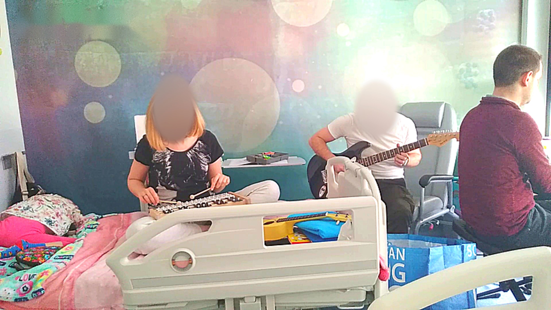 Sala szpitalna: osoba na łóżku gra na cymbałkach, mężczyzna obok na gitarze, Michał Drezner przy oknie, twarze pozostałych zasłonięte