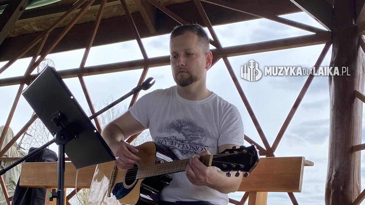 Michał Drezner gra na gitarze w drewnianej altanie, obok statyw z nutami