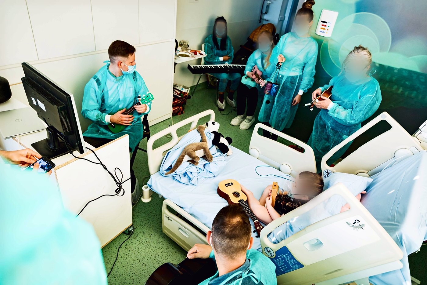 Sala szpitalna z monitorem i łóżkiem, kilkoro grających w fartuchach, dziecko z ukulele